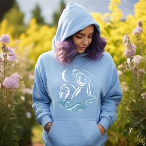 Puede incluir: Una sudadera con capucha azul claro con un gr&aacute;fico blanco de dos caballos corriendo a trav&eacute;s de olas con una luna creciente y estrellas en el fondo.