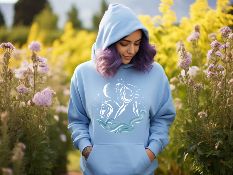 Puede incluir: Una sudadera con capucha azul claro con un gr&aacute;fico blanco de dos caballos corriendo a trav&eacute;s de olas con una luna creciente y estrellas en el fondo.