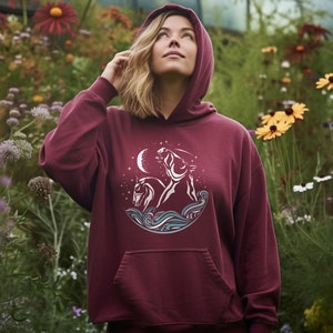 Puede incluir: Una sudadera con capucha color burdeos con un gr&aacute;fico blanco de dos caballos corriendo por las olas bajo una luna creciente y estrellas.