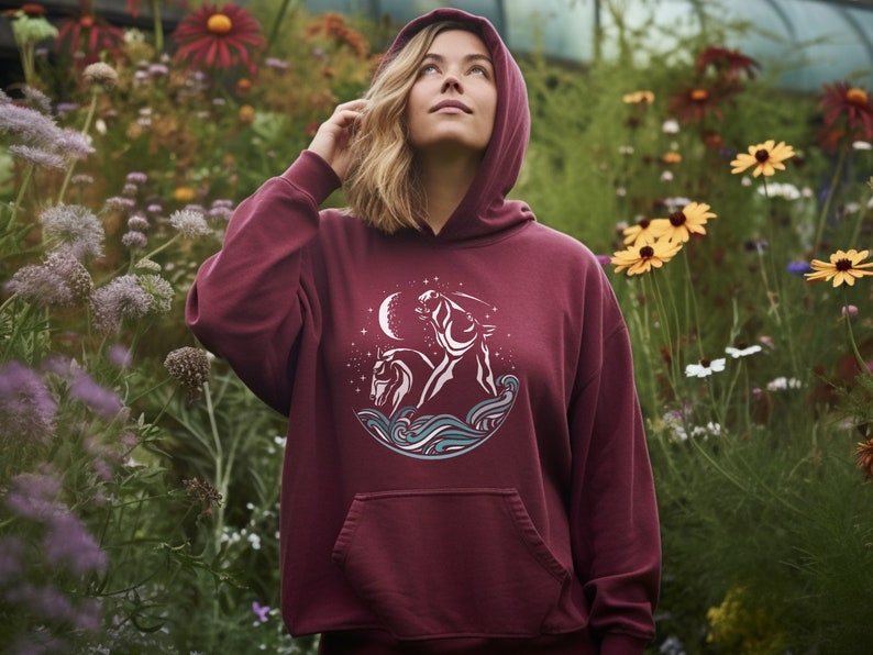 Puede incluir: Una sudadera con capucha color burdeos con un gr&aacute;fico blanco de dos caballos corriendo por las olas bajo una luna creciente y estrellas.