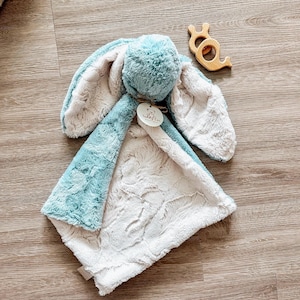 以下が含まれることがあります： 青とクリーム色のベビーブランケットとウサギのおもちゃ。ウサギは長い耳と柔らかい質感を持っています。木製のカタツムリ型歯固めも写っています。