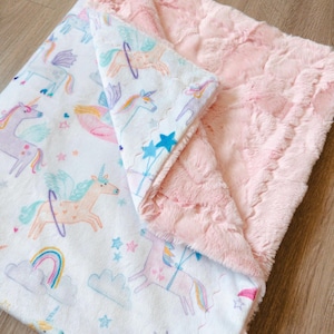 Può includere: Coperta per bambini rosa e bianca con tema unicorno. La coperta presenta un lato bianco con unicorni colorati, arcobaleni e stelle e un lato posteriore rosa, in peluche e strutturato.