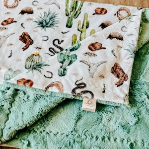 May include: A soft, plush baby blanket with a white top featuring a Southwestern design of cacti, boots, and a snake. The underside is a solid, textured, mint green. The blanket is folded, showing both sides.