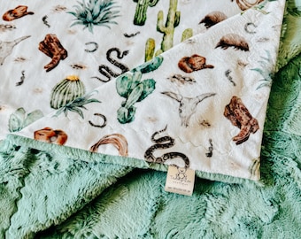 May include: A soft, plush baby blanket with a white top featuring a Southwestern design of cacti, boots, and a snake. The underside is a solid, textured, mint green. The blanket is folded, showing both sides.
