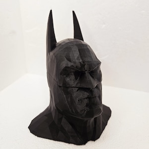 Op de afbeelding: Een zwarte, geometrische Batman-buste, met het masker en de puntige oren van het personage. De sculptuur is 3D-geprint, met zichtbare laaglijnen die een facetachtig uiterlijk creëren. De buste staat tegen een effen witte achtergrond.