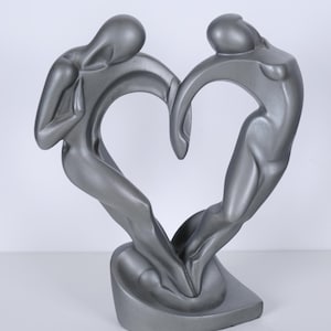 May include: A metallic silver sculpture depicting two stylized human figures leaning toward each other to form a heart shape. The figures are connected at the base and reach upward, with their curved bodies creating an open space in the center.