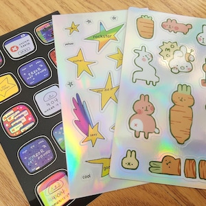 May include: Three sheets of holographic stickers with various designs. The first sheet features a rainbow-colored shooting star, a yellow star with the word "mother", a pink star with the word "rockstar", and a blue star with the word "yahoo". The second sheet features a variety of stickers with text including "404 where is it", "error not found", "my other is broken", "signal", "system error", and "cool". The third sheet features a variety of cartoon stickers including a carrot, a bunny, a llama, and a variety of other animals.