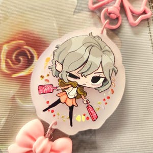 May include: A round, clear keychain with a colorful illustration of a cartoon character holding two pink objects. The character has gray hair and is wearing a brown jacket and a pink skirt. The keychain is decorated with glitter and small, colorful dots.
