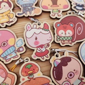 May include: A collection of colorful wooden charms featuring various cartoon characters. The charms are attached to keychains with a small metal ring. The characters include a dog, a cat, a squirrel, a bunny, a frog, a mushroom, and a snail.