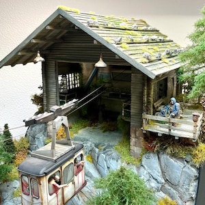 Könnte beinhalten: Ein Modell einer kleinen Holzhütte mit grünem Dach und einer Seilbahn, die an einem Draht hängt. Die Hütte hat eine kleine Veranda mit einem Geländer und eine Figur, die auf einer Bank sitzt. Die Seilbahn ist weiß mit roten Akzenten und hat zwei Figuren im Inneren.
