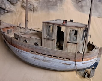 May include: A weathered wooden model of a small fishing boat with a white painted hull and a brown wooden cabin. The boat is sitting on a sandy surface.