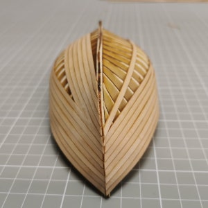 以下が含まれることがあります： 木製の模型船の船体で、細かい板張りの模様が施されています。船体は薄い木片を接着して、滑らかで湾曲した表面に仕上げられています。