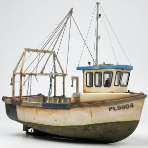 May include: A scale model fishing trawler with a weathered cream hull, a blue roof cabin, and a tall mast structure. The side of the hull features the black text PL9904. The vessel includes a rear gantry, rigging lines, and a green textured waterline.