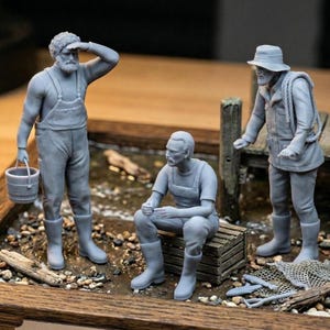 May include: A detailed miniature diorama featuring three grey-toned figures of fishermen. One figure holds a bucket, another shields his eyes, and the third leans against a wooden post. The scene includes a small body of water and fishing gear.
