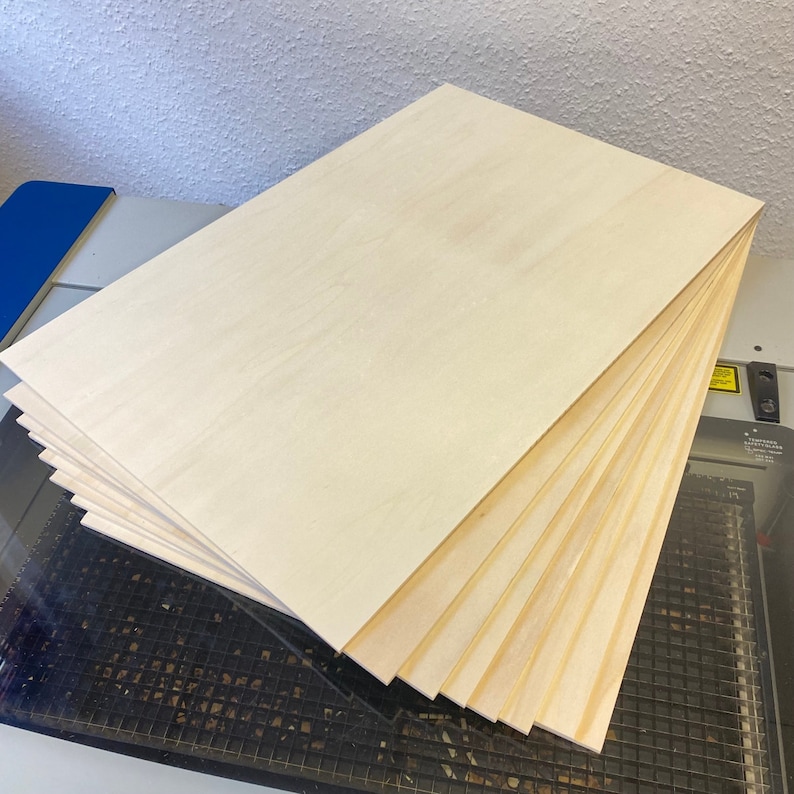 May include: A stack of thin, light wood panels, likely plywood, with a smooth, light-colored surface. The panels are stacked on top of each other, with the top panel being the largest. The panels are on a black surface with a grid pattern.