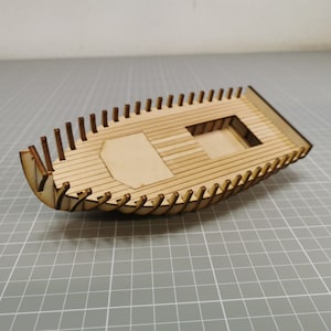 以下が含まれることがあります： 詳細な船体と甲板を持つ木製模型船キット。キットは薄い茶色の木で作られており、長方形です。船には甲板に小さな開口部があり、これはキャビンまたはその他の機能用です。