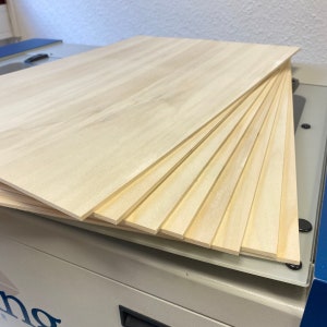 May include: A stack of thin, light wood boards, likely plywood, with a natural wood grain finish. The boards are stacked on top of each other, with the top board slightly angled. The boards are on a white surface.