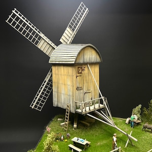 May include: A detailed miniature model of a wooden windmill with four blades. The windmill has a weathered, aged appearance with a shingled roof. A small picnic table and figures are in the foreground, set on a grassy base. The model includes a ladder and a small wooden balcony.