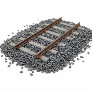 Può includere: Una sezione di binario ferroviario in miniatura con due rotaie metalliche parallele color ruggine fissate su traverse in legno grigio usurato. L'assemblaggio del binario è inserito in un letto di pietrisco grigio scuro.
