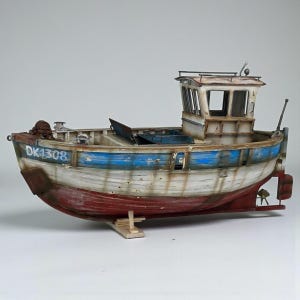 Könnte beinhalten: Detailliertes Modell eines Fischerbootes mit verwittertem Aussehen in Weiß, Blau und Rot. Das Boot hat eine Kabine mit Fenstern und den Text "DK1308" am Bug. Das Modell steht vor einem schlichten Hintergrund.
