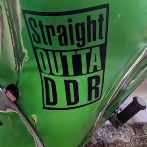Könnte beinhalten: Grüne Fahrzeugverkleidung mit dem Text "Straight Outta DDR" in Schwarz und Weiß. Die Verkleidung weist Gebrauchsspuren auf, mit Kratzern und Unvollkommenheiten. Ein Lenker und andere Fahrzeugteile sind sichtbar.