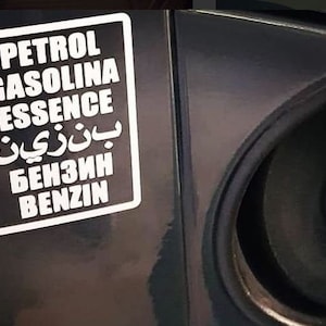 Puede incluir: Una pegatina rectangular en blanco y negro con las palabras "PETROL GASOLINA ESSENCE" en inglés, árabe, ruso y alemán. Se encuentra adherida a una superficie oscura, cerca de la tapa del depósito de combustible.