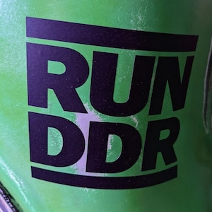 Op de afbeelding: Close-up van een groene ondergrond met de zwarte tekst "RUN DDR" in een vetgedrukte, schreefloze lettertype. De letters zijn gestapeld, met "RUN" boven "DDR". De achtergrond is effen, heldergroen.
