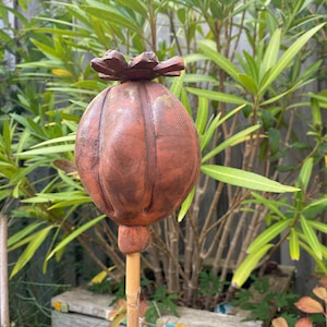 Puede incluir: Una estaca de jardín decorativa con forma de cabeza de semilla de amapola de color óxido. La cabeza de la semilla es redonda con surcos verticales y una parte superior en forma de flor. La estaca es de bambú y está sobre un fondo de follaje verde.