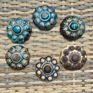 May include: Six decorative ceramic knobs in shades of teal, blue, and brown. Each knob features a central circular design surrounded by smaller, textured details. The knobs are arranged on a woven, neutral-toned surface.