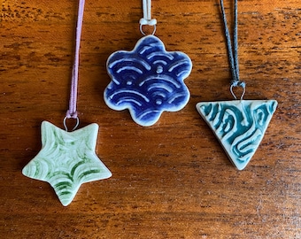 May include: Three ceramic pendants on cords. The pendants are a blue flower with a wave pattern, a green star with a swirl pattern, and a green and blue triangle with a swirl pattern.