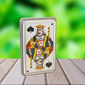 May include: A deck of playing cards featuring the King of Spades. The card has a detailed illustration of a king in a crown, holding a scepter. The card is encased in a clear plastic case, set against a blurred green background.