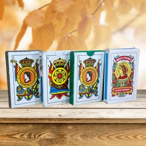 Könnte beinhalten: Vier Vintage-Spielkartensets mit aufwendigen Designs und Text, darunter die Wörter "MARIA ESPAÑA" und "CALIDAD SUPERIOR". Die Decks zeigen königliche Bilder und werden auf einer Holzoberfläche vor einem verschwommenen Herbsthintergrund präsentiert.