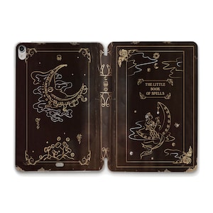 May include: A black leather-bound book with gold accents and illustrations. The front cover features a crescent moon with a face and the text "The Little Book of Spells". The back cover features a mermaid sitting on a crescent moon.