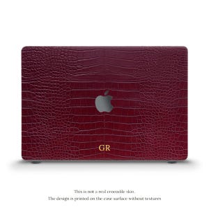May include: A burgundy laptop case with a faux crocodile skin texture. The case features the Apple logo and the initials "GR" in gold. The text at the bottom reads, "This is not a real crocodile skin. The design is printed on the case surface without textures."