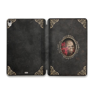 Puede incluir: Una funda de iPad de cuero negro con un diseño dorado ornamentado. La cubierta frontal presenta una calavera con flores rojas en un marco dorado.