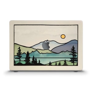 May include: A laptop skin with a watercolor design of a mountain range with a lake in the foreground. The mountains are blue, green, and purple, and the lake is blue. There are trees in the foreground and a yellow sun in the sky.