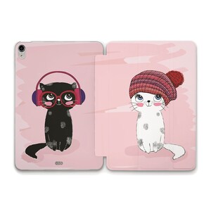May include: A pink iPad case featuring a black cat wearing headphones and red glasses on the left side and a white cat wearing a red and pink knitted hat on the right side.