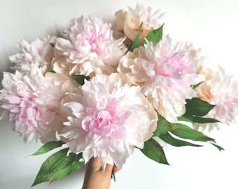 May include: A bouquet of light pink and cream-colored peonies with green leaves. The flowers have multiple layers of delicate petals, with a deeper pink hue in the center. The bouquet is held in a hand.