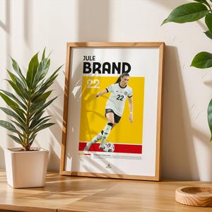 Może przedstawiać: Oprawiony plakat z piłkarką w biało-czarnym stroju z numerem 22. Plakat ma ż&oacute;łto-czerwone tło z napisem "Jule Brand" u g&oacute;ry. Plakat stoi na drewnianym stole obok rośliny doniczkowej.