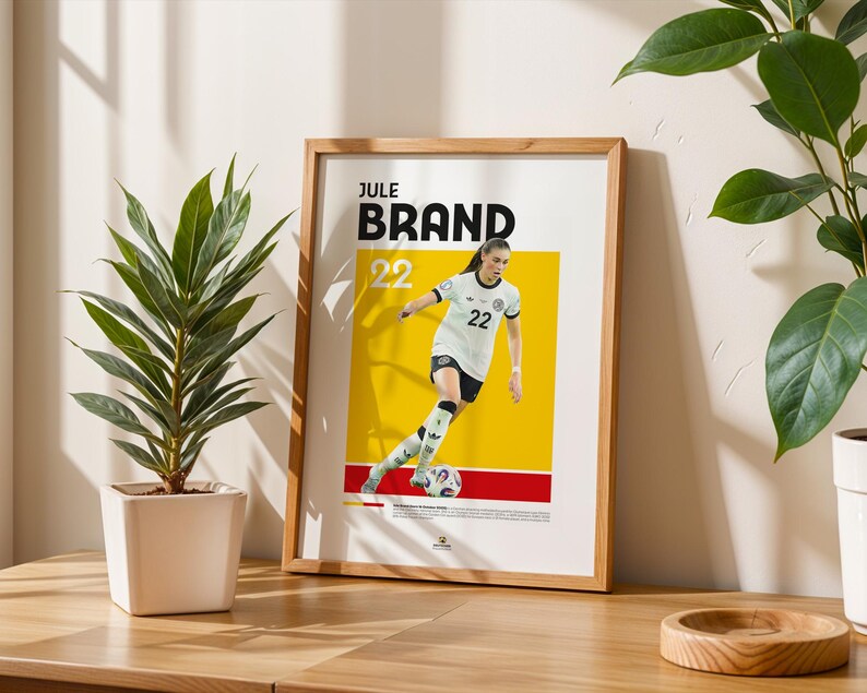 Może przedstawiać: Oprawiony plakat z piłkarką w biało-czarnym stroju z numerem 22. Plakat ma ż&oacute;łto-czerwone tło z napisem "Jule Brand" u g&oacute;ry. Plakat stoi na drewnianym stole obok rośliny doniczkowej.