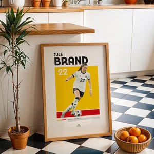 Może przedstawiać: Oprawiony plakat z nazwiskiem "JULE BRAND" i numerem "22", przedstawiający piłkarkę w biało-czarnym stroju na ż&oacute;łtym tle. Widoczna jest r&oacute;wnież mała roślina doniczkowa i kosz pomarańczy.