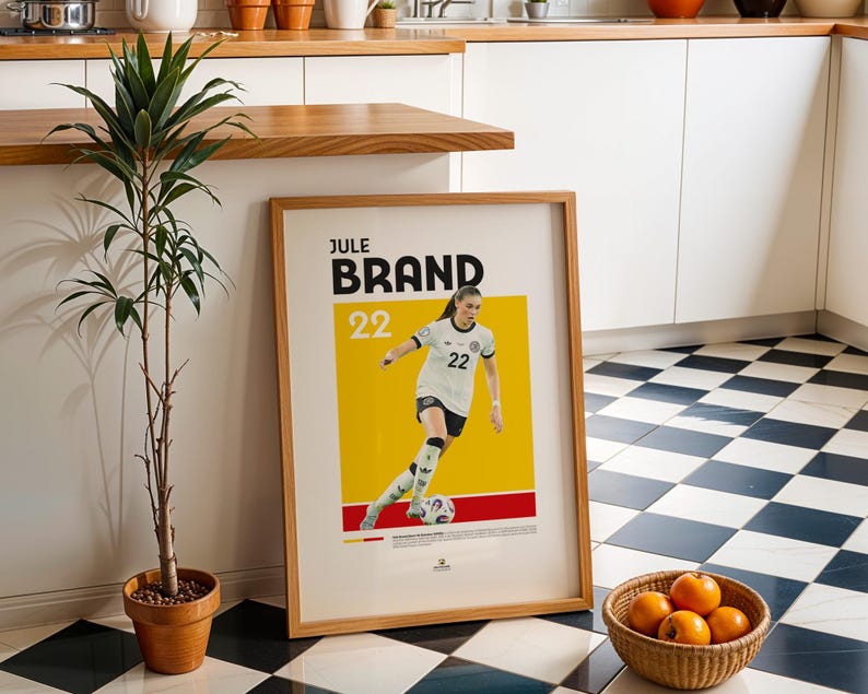 Może przedstawiać: Oprawiony plakat z nazwiskiem "JULE BRAND" i numerem "22", przedstawiający piłkarkę w biało-czarnym stroju na ż&oacute;łtym tle. Widoczna jest r&oacute;wnież mała roślina doniczkowa i kosz pomarańczy.