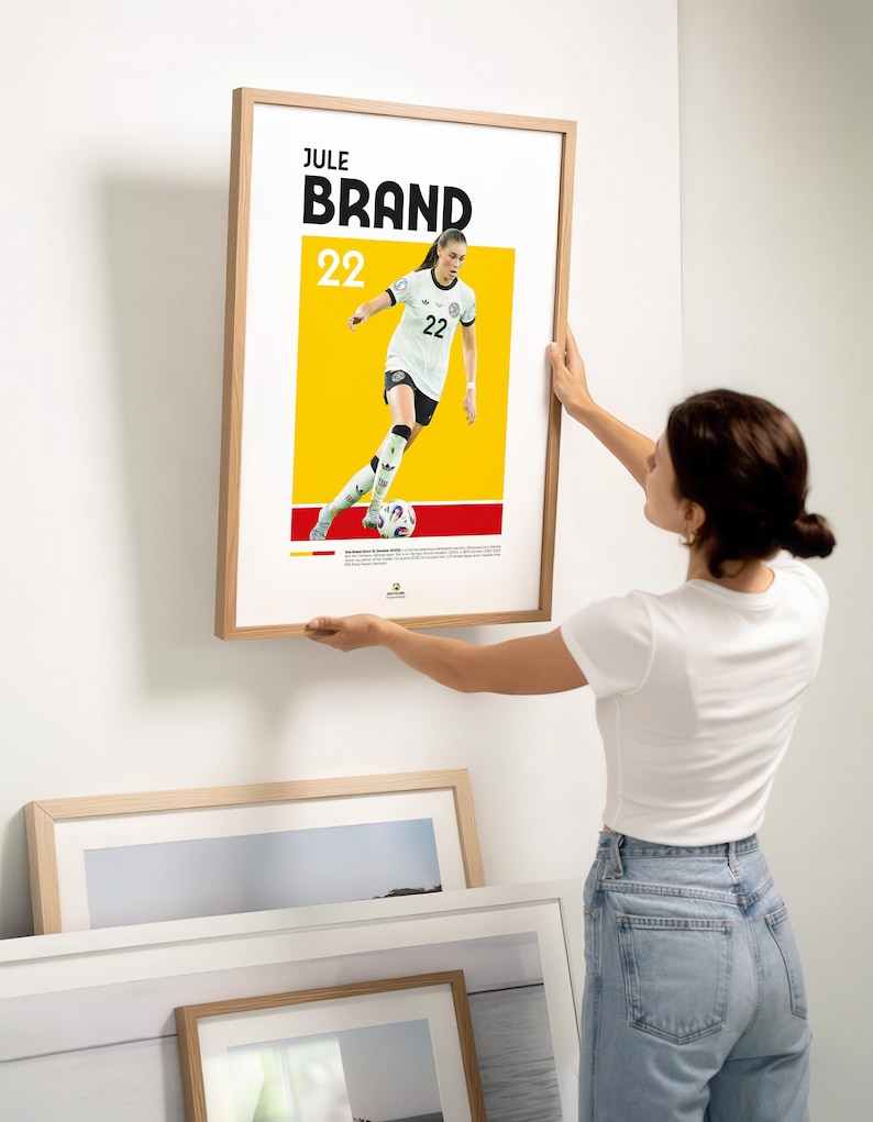 Może przedstawiać: Oprawiony plakat z Jule Brand, numer 22, w stroju piłkarskim. Plakat ma białe tło z ż&oacute;łto-czerwonym paskiem. Plakat jest trzymany przez osobę.