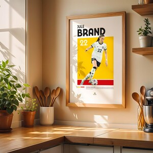 Może przedstawiać: Oprawiony plakat z piłkarką w biało-czarnym stroju, z nazwiskiem "JULE BRAND" i numerem "22" na pierwszym planie. Tło jest ż&oacute;łte i czerwone. Plakat jest w drewnianej ramie.