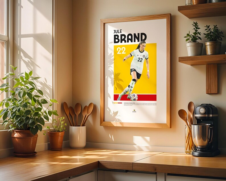 Może przedstawiać: Oprawiony plakat z piłkarką w biało-czarnym stroju, z nazwiskiem "JULE BRAND" i numerem "22" na pierwszym planie. Tło jest ż&oacute;łte i czerwone. Plakat jest w drewnianej ramie.