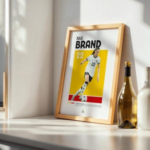 Może przedstawiać: Oprawiony plakat z piłkarką w biało-czarnym stroju, z nazwiskiem "Jule Brand" i numerem "22". Plakat ma ż&oacute;łto-czerwone tło. Obok plakatu stoi butelka wina i biała butelka.