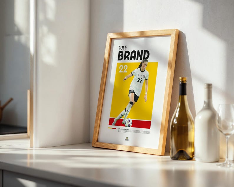 Może przedstawiać: Oprawiony plakat z piłkarką w biało-czarnym stroju, z nazwiskiem "Jule Brand" i numerem "22". Plakat ma ż&oacute;łto-czerwone tło. Obok plakatu stoi butelka wina i biała butelka.