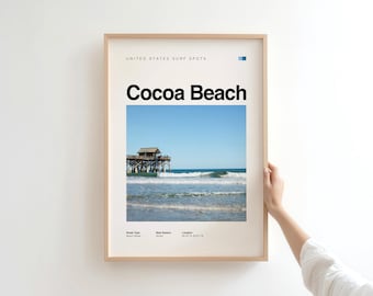 May include: Framed print featuring "Cocoa Beach" in bold black text over a beach scene. The image shows a pier extending into the ocean under a clear blue sky. The print is in a light wood frame, held by a person.