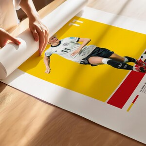 May include: A poster depicting a soccer player in a white and black kit with the number 11, set against a yellow and red backdrop. A football is near the player's feet. The poster is being unrolled.
