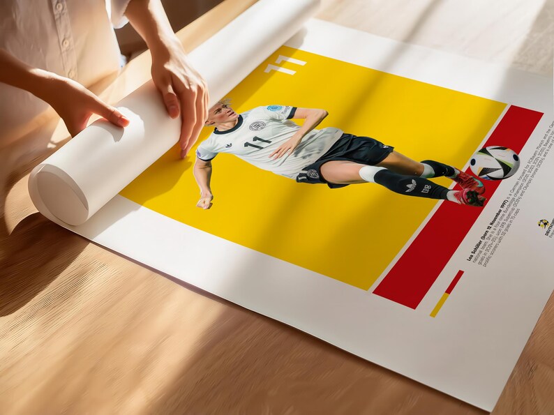 May include: A poster depicting a soccer player in a white and black kit with the number 11, set against a yellow and red backdrop. A football is near the player's feet. The poster is being unrolled.